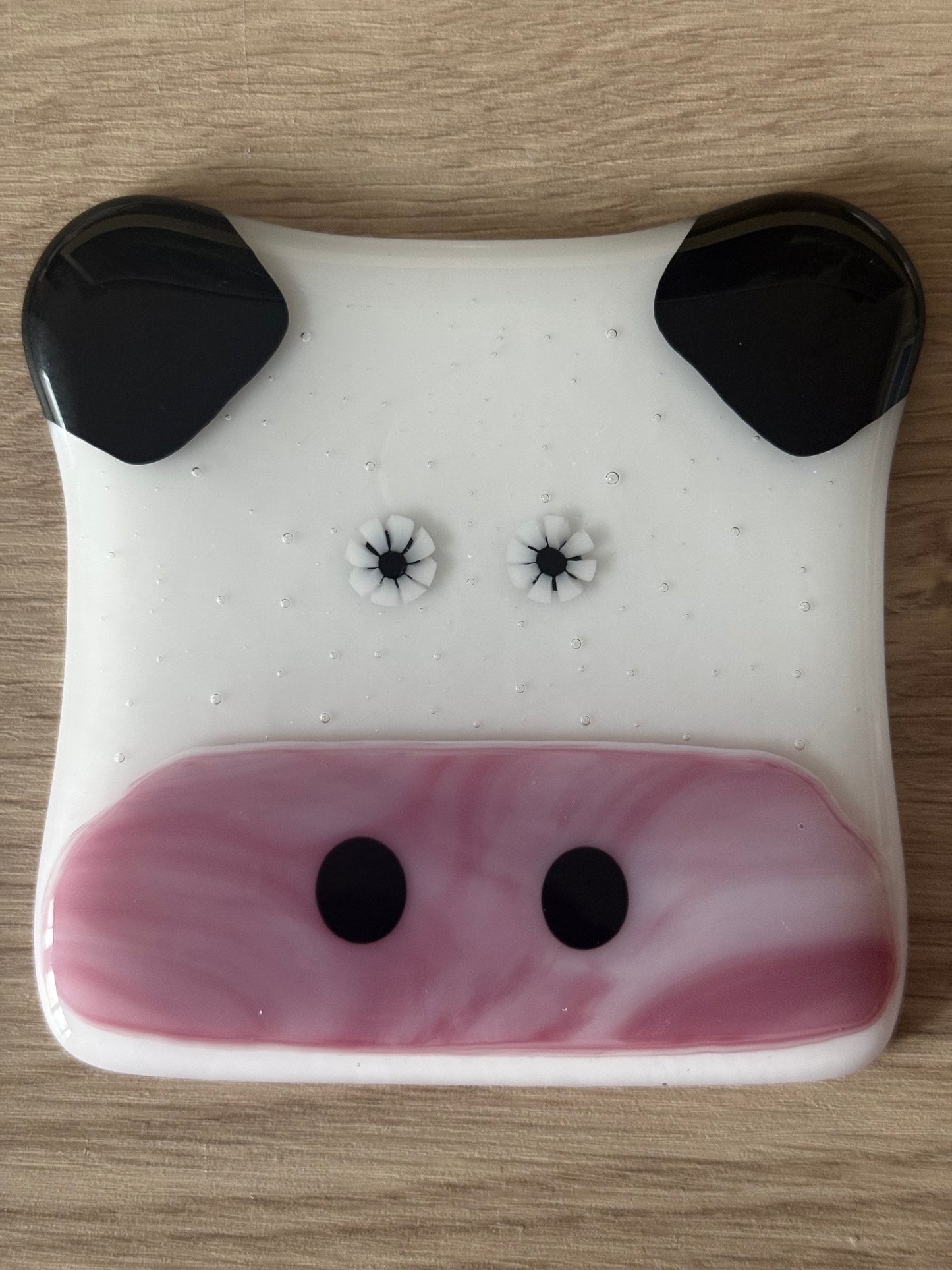 A handmade fused glass coaster designed to resemble a cow with a white body, black ears, and pink details.
