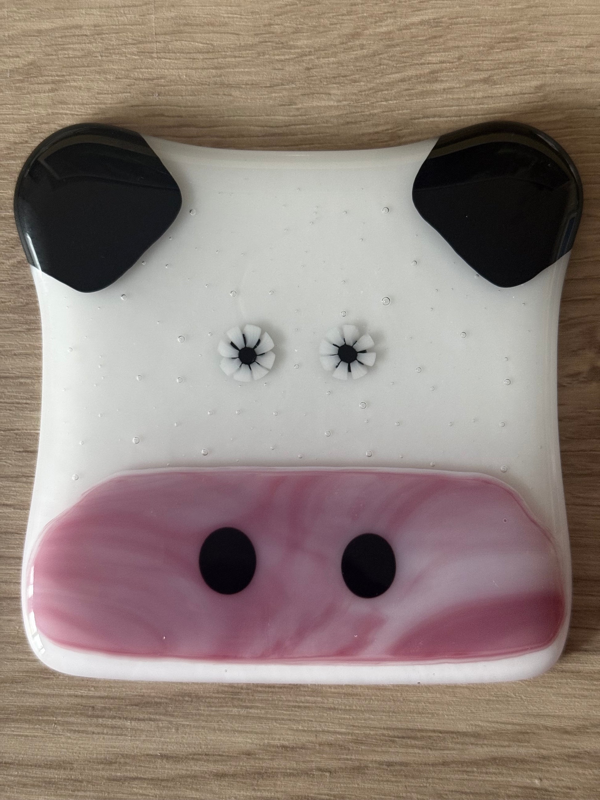A handmade fused glass coaster designed to resemble a cow with a white body, black ears, and pink details.